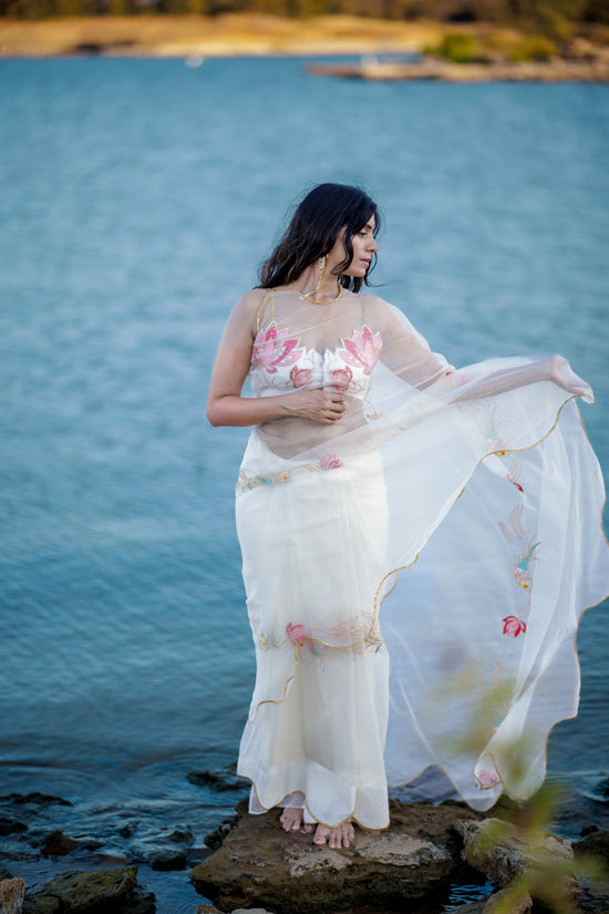 A woman stands barefoot on rocks by the water, wearing a flowing sheer white organza saree with pink fish sequin embroidery with a wave patterned pitta embroidery border