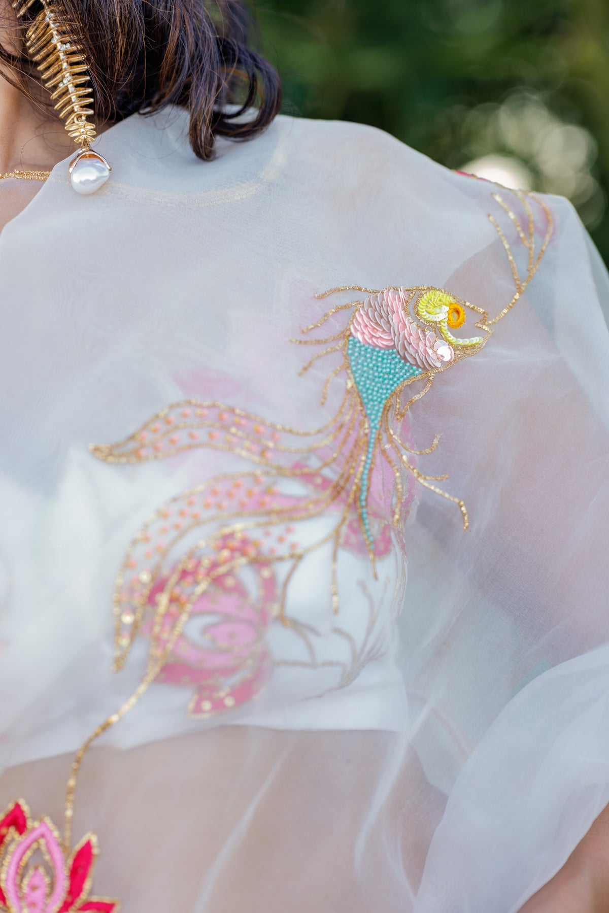 A close-up of a white sheer organza saree featuring an intricate, colorful embroidered fish design with pink, teal, yellow, and gold details.