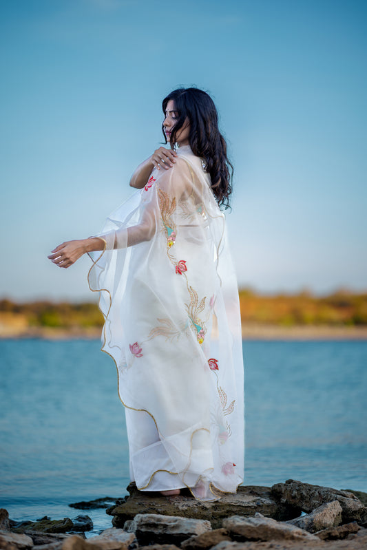 A woman wearing a flowing white embroidered pure organza saree stands on rocks by the water, with blue sky and distant shoreline in the background.