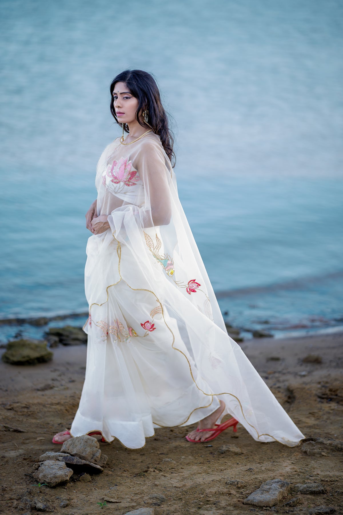 A woman wearing a flowing white embroidered sari stands on a sandy shore beside the water. The garment has pink sequinembroidery and gold trim, with sheer sleeves and a long draped fabric.