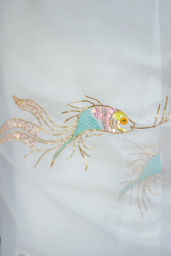 Close-up of a white embroidered organza saree with delicate fish and lotus motifs in pink, teal, and gold thread, shown against a blurred green outdoor background.