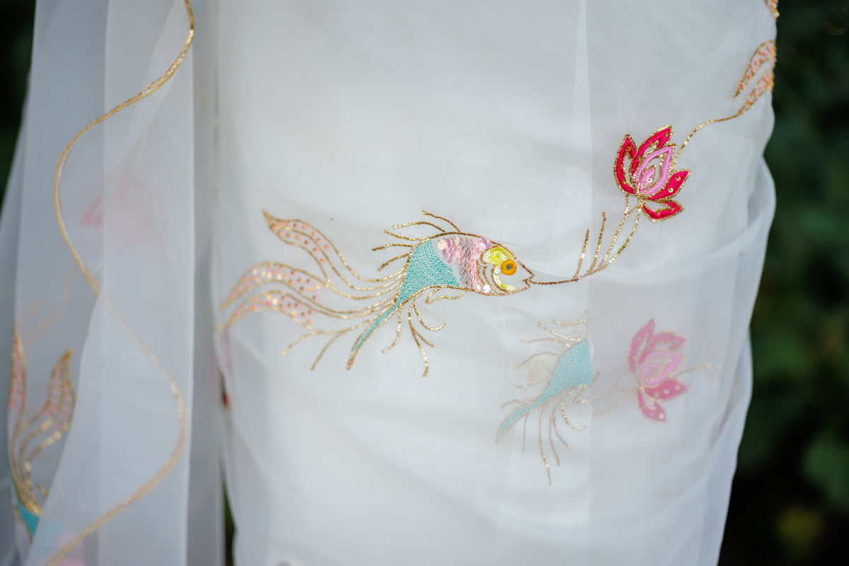Close-up of a white embroidered organza saree with delicate fish and lotus motifs in pink, teal, and gold thread, shown against a blurred green outdoor background.