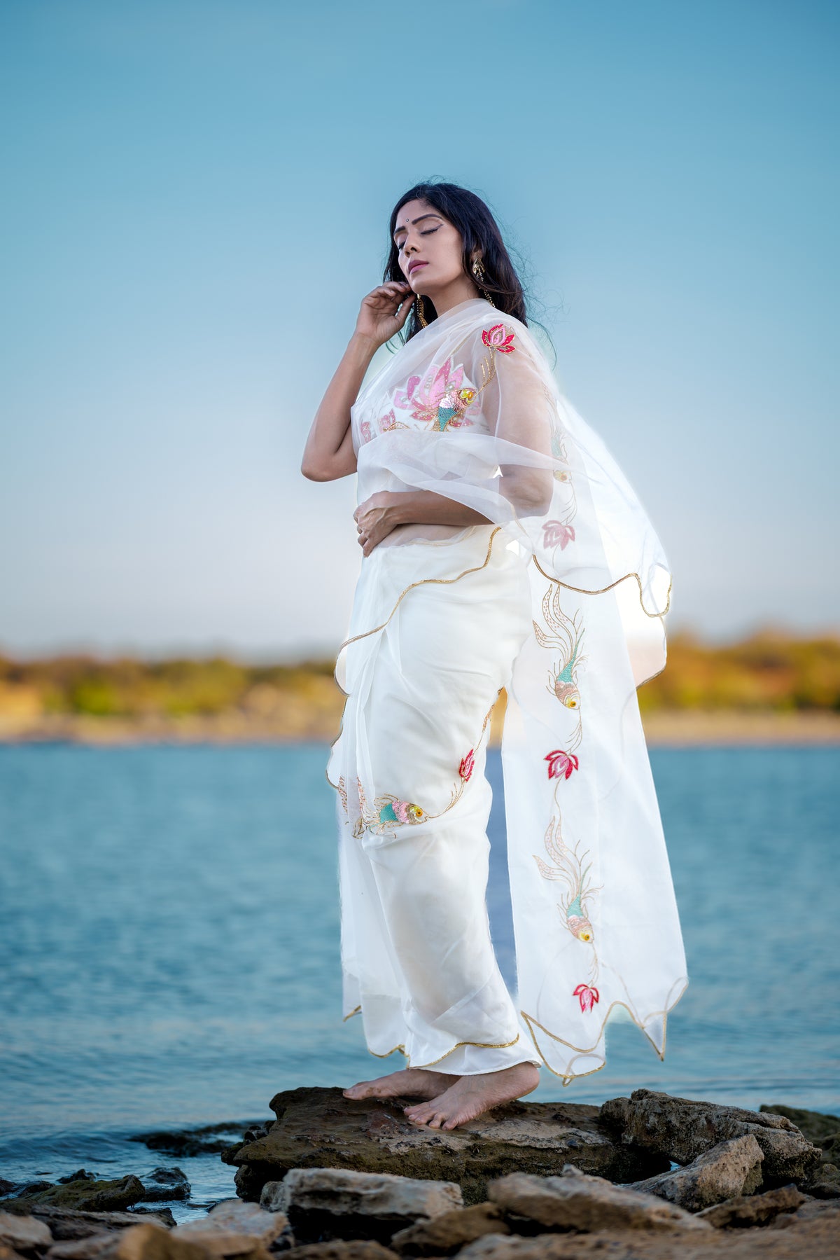A woman stands barefoot on rocks by the water wearing a white embroidered saree with floral and bird motifs, against a blue sky and sea background.