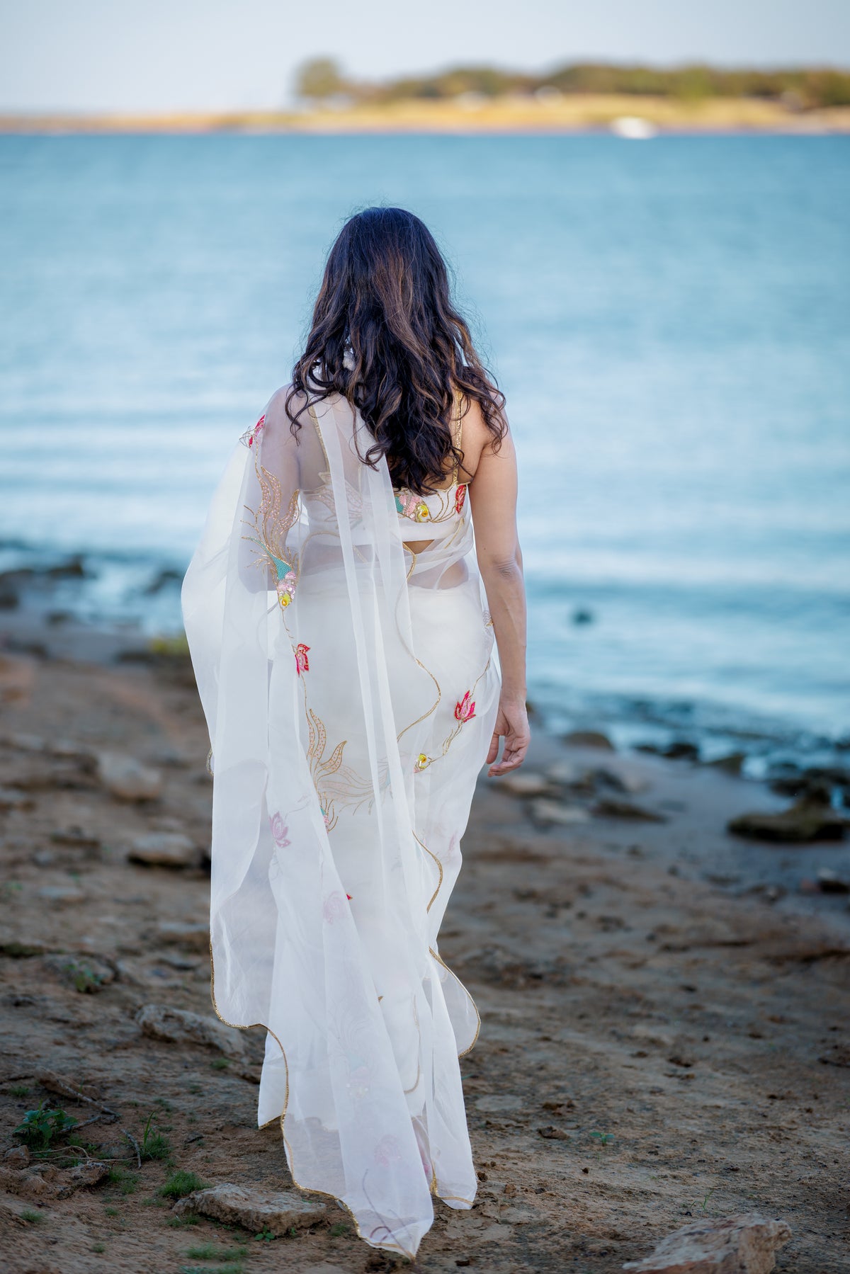 A woman viewed from behind walks along a rocky shoreline toward the ocean, wearing a flowing white saree with delicate sequin and pitta embroidery 