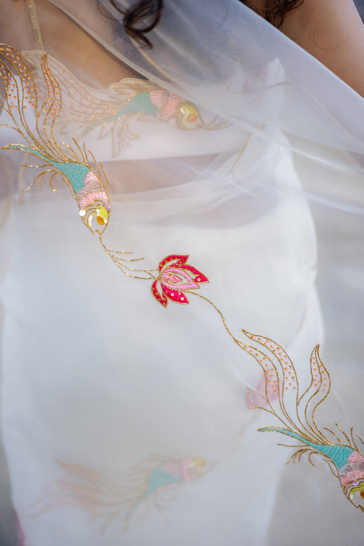 Close-up of a white sheer embroidered saree, featuring gold threadwork and colorful floral motifs in pink, teal, and yellow.