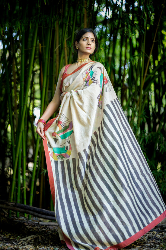 A woman wearing a traditional Indian kalamkari saree stands in front of a dense bamboo forest. The saree has a powder pink cream-colored body with black stripes, a red border, and colorful floral prints on the pallu.