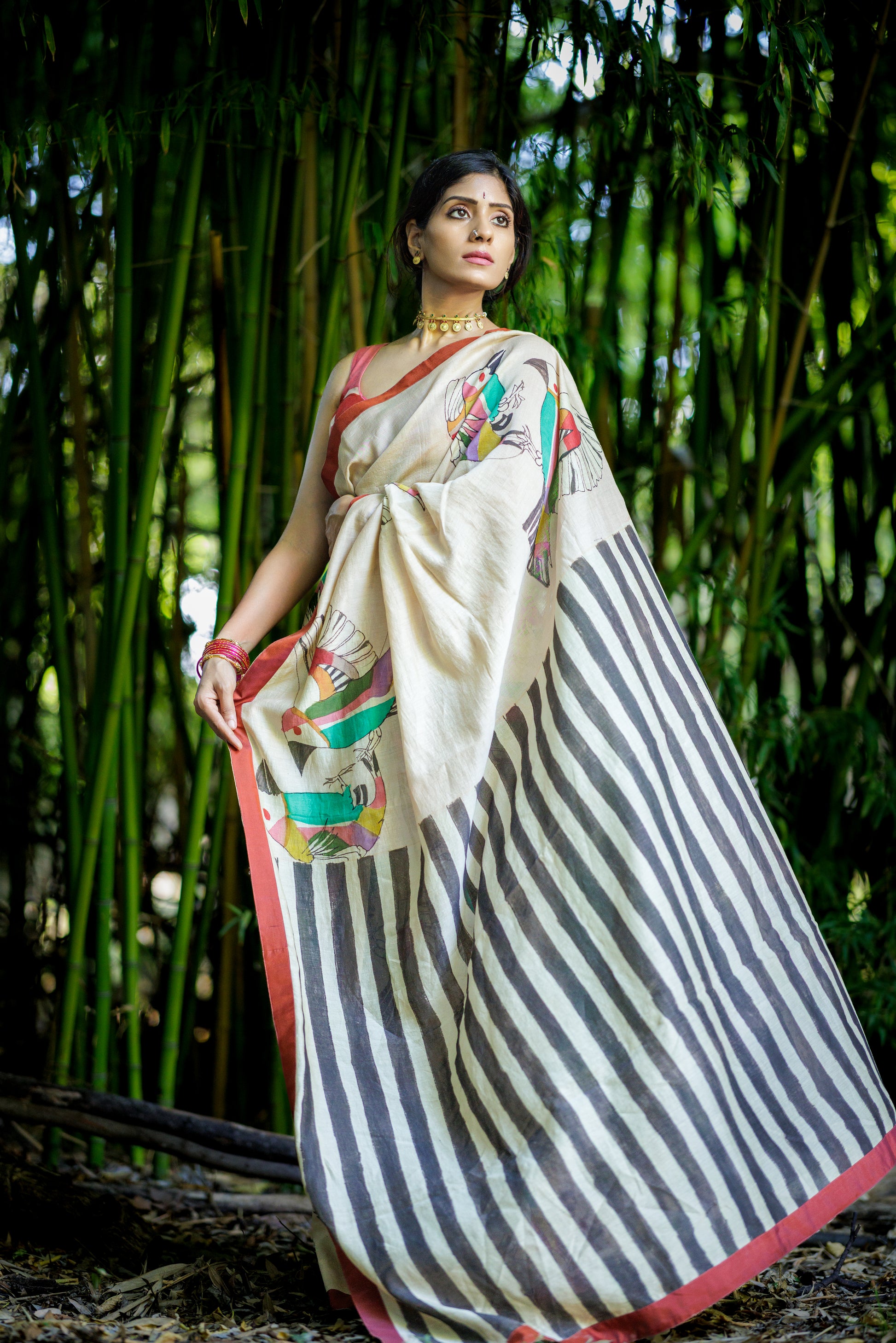 A woman wearing a traditional Indian kalamkari saree stands in front of a dense bamboo forest. The saree has a powder pink cream-colored body with black stripes, a red border, and colorful floral prints on the pallu.