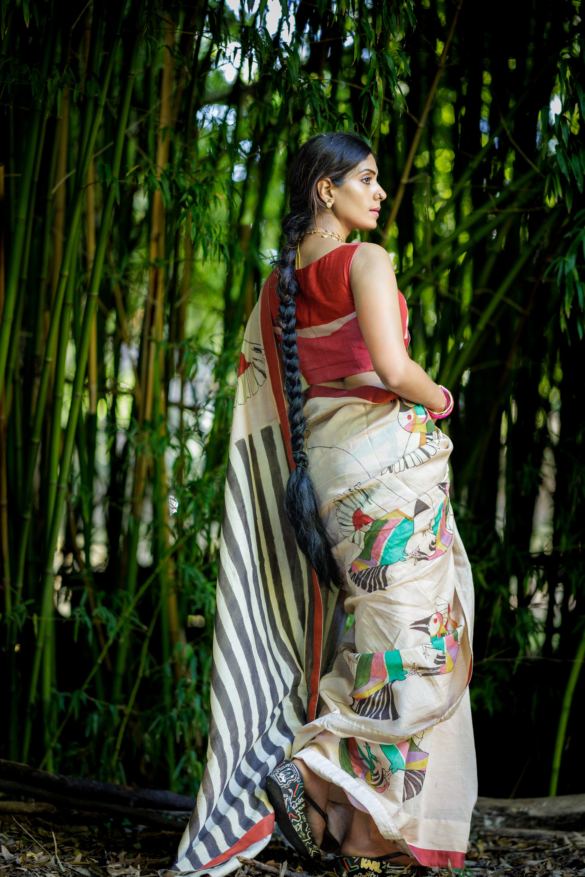 A woman wearing a traditional Indian kalamkari saree stands in front of a dense bamboo forest. The saree has a beige base with black stripes on the pallu and colorful hand-painted motifs, paired with a red sleeveless blouse.