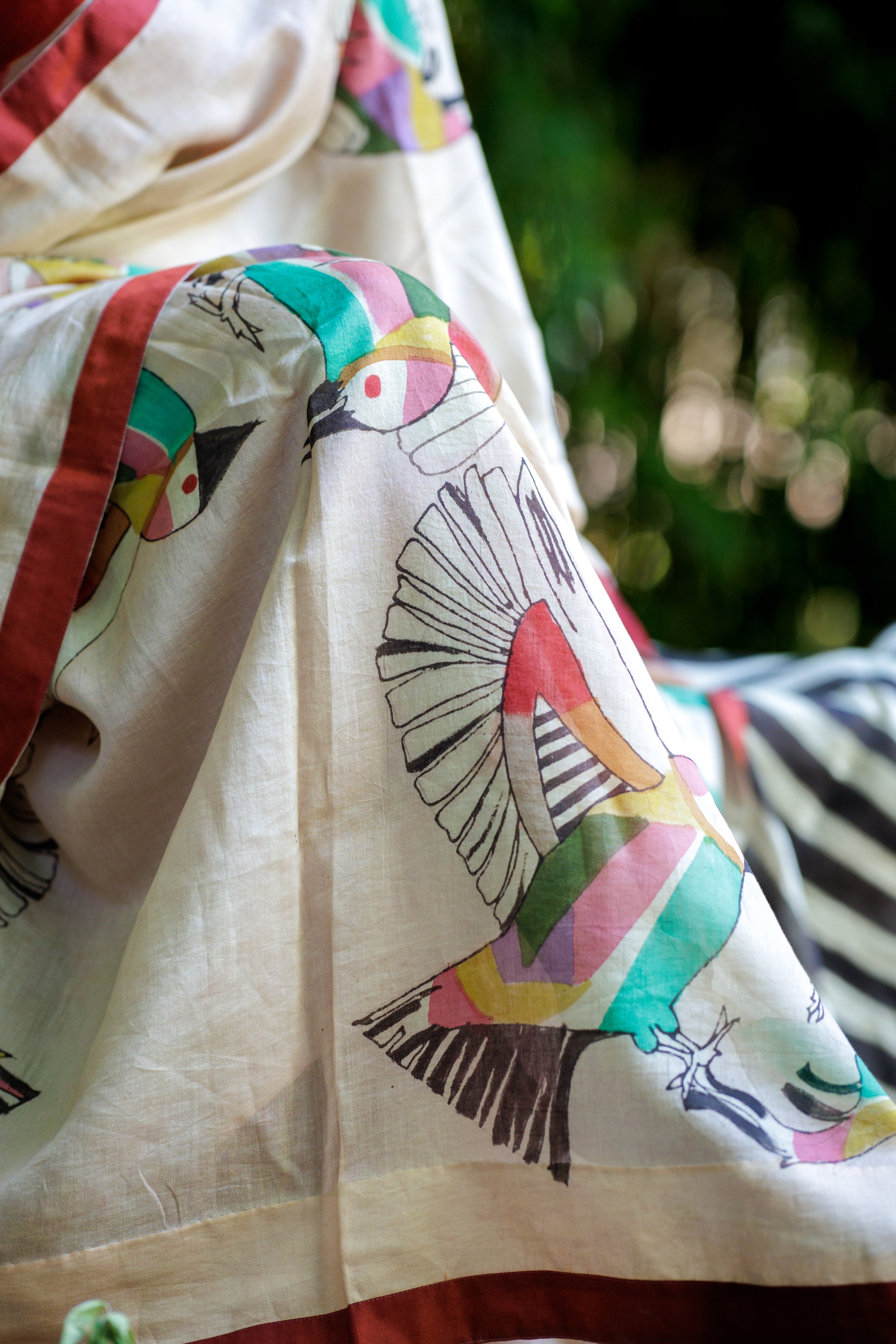 A close-up of a draped cream-colored fabric with a colorful hand-painted bird pattern and a red border, set against a blurred green outdoor background.