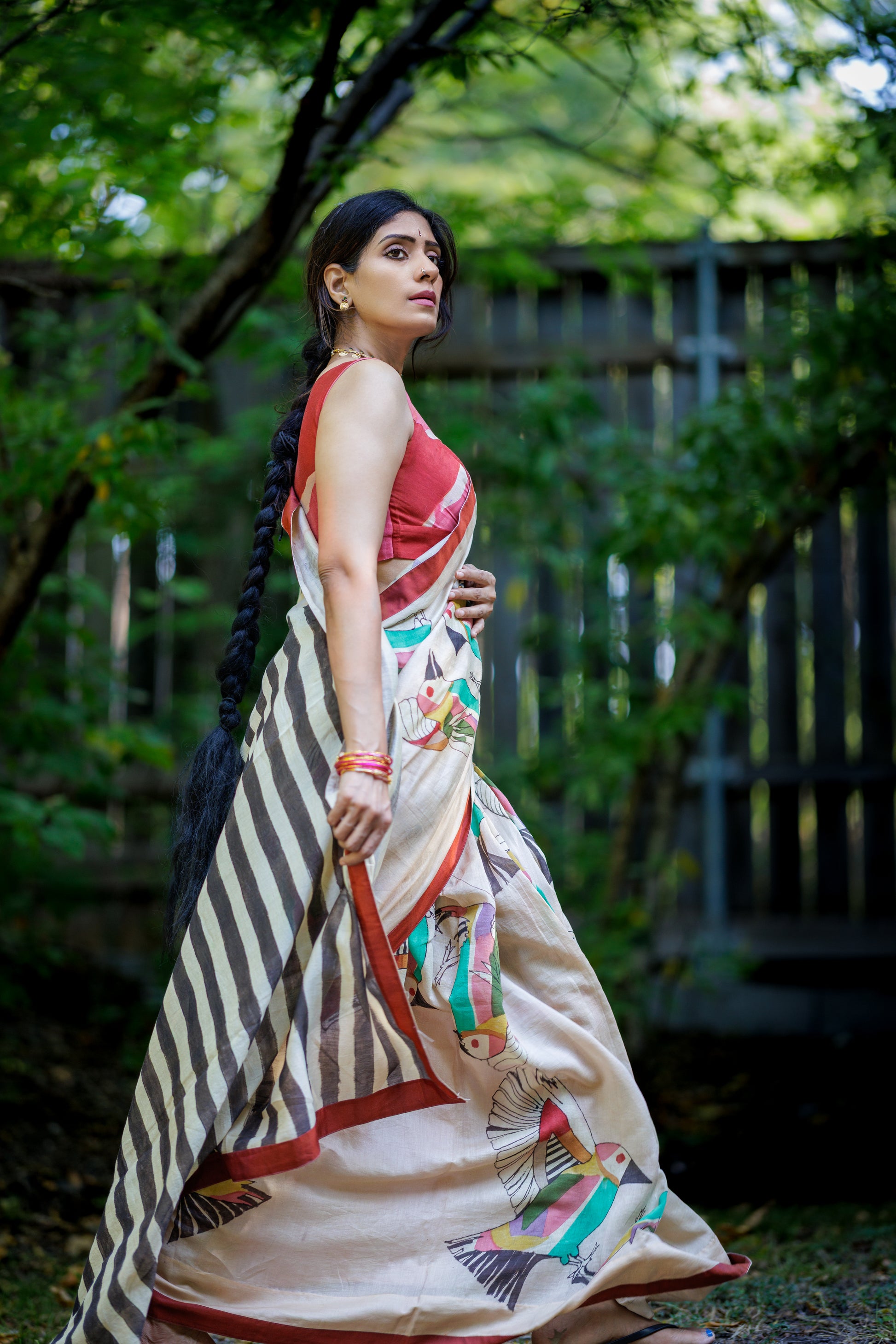 A woman wearing a traditional Indian saree stands outdoors in a garden setting. The saree has a beige base with black stripes on one side, a red sleeveless blouse, and colorful printed motifs near the hem.