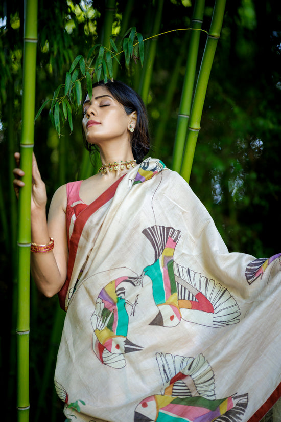 A woman wearing a powder pink colored saree with colorful bird prints, posed in front of a lush green bamboo forest.