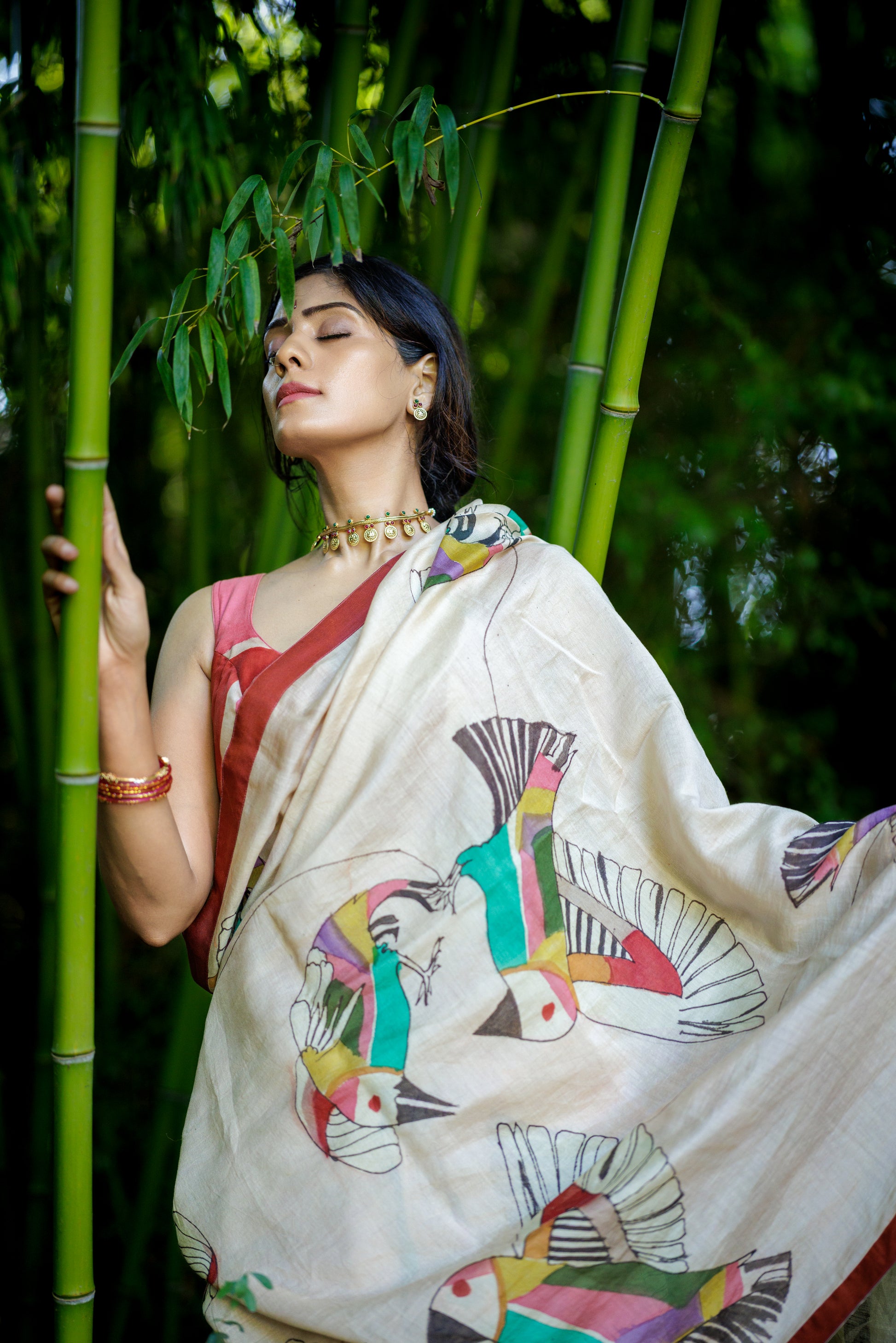 A woman wearing a powder pink colored saree with colorful bird prints, posed in front of a lush green bamboo forest.