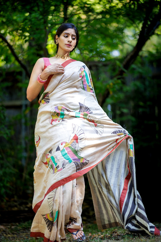 A woman outdoors wearing a cream-colored hand-painted saree with colorful  abstract bird motifs and a red border, standing in a wooded setting.