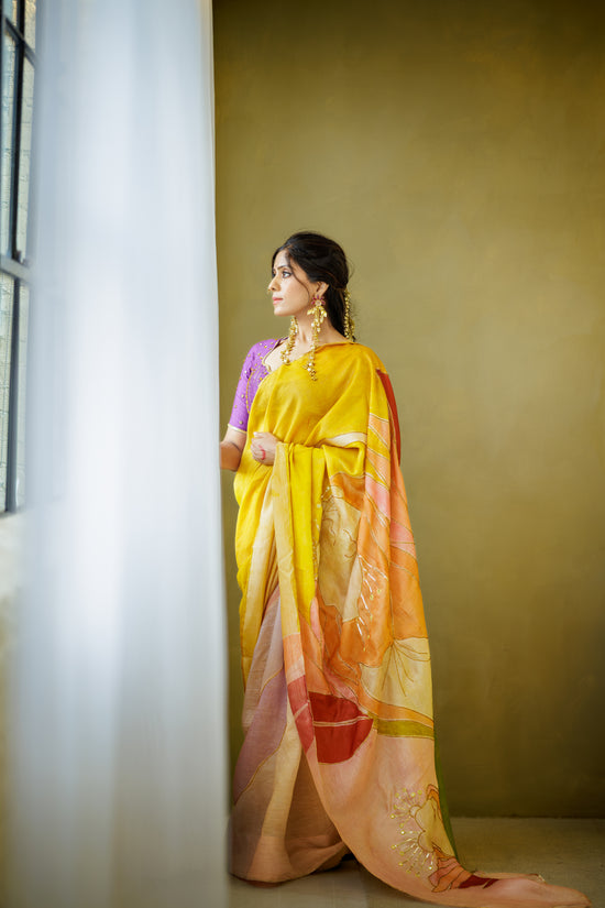 A woman wearing a yellow and orange saree with a purple blouse stands beside a window in a room with a textured mustard wall.