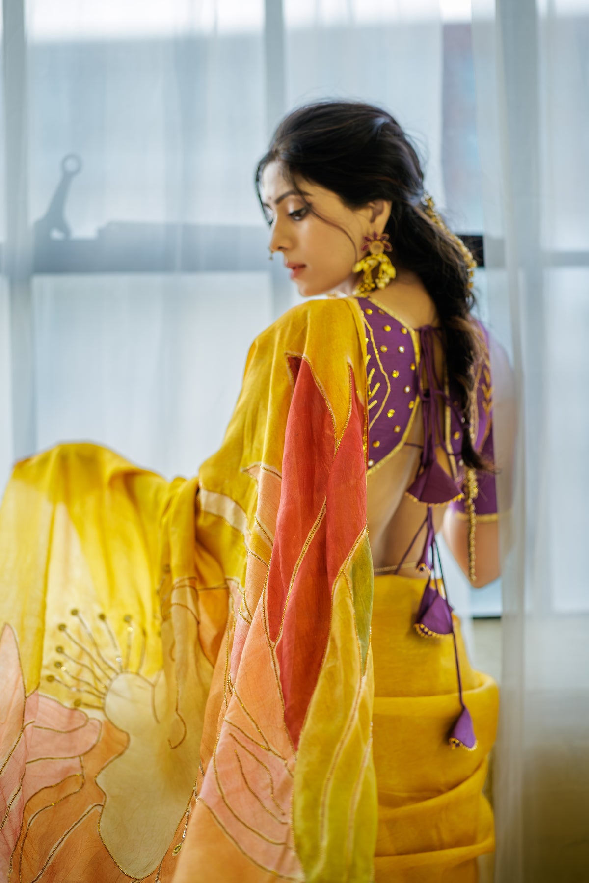 A woman wearing a vibrant yellow saree with orange and pink floral patterns, paired with a purple blouse featuring gold polka dots and tie-back details, standing in front of a softly lit window.