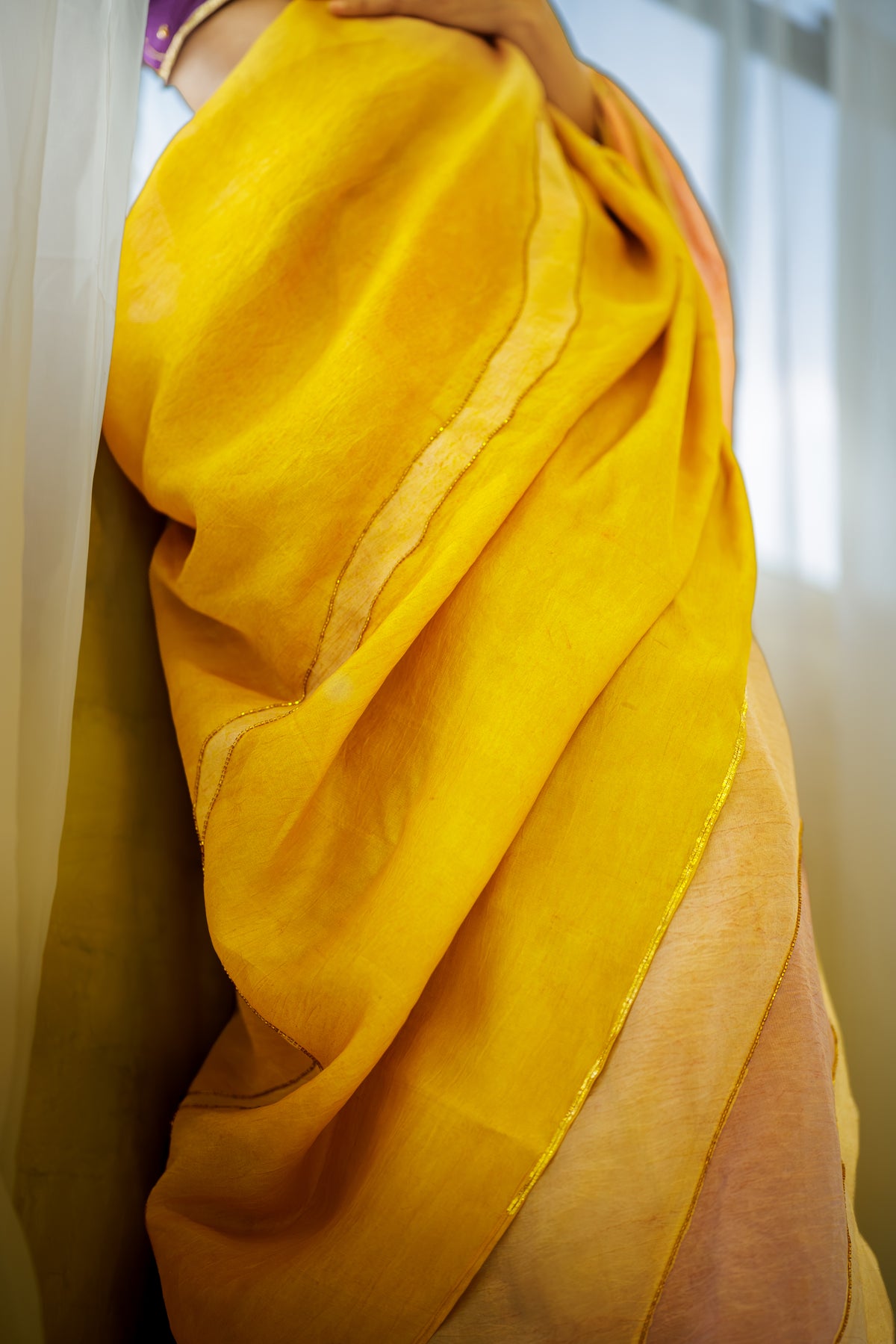 A close-up of a person wearing a draped mustard yellow fabric, likely a sari, with soft folds and subtle metallic border details.