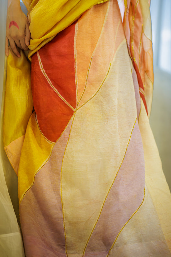 A close-up of a lightweight draped fabric or sari with large abstract leaf-like panels in yellow, orange, blush pink, and cream, outlined with thin gold lines. A person's hands are visible holding the fabric.