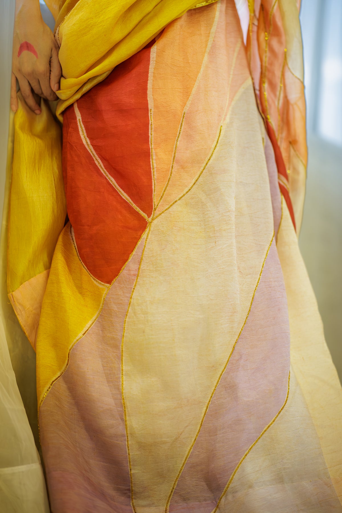 A close-up of a lightweight draped fabric or sari with large abstract leaf-like panels in yellow, orange, blush pink, and cream, outlined with thin gold lines. A person's hands are visible holding the fabric.