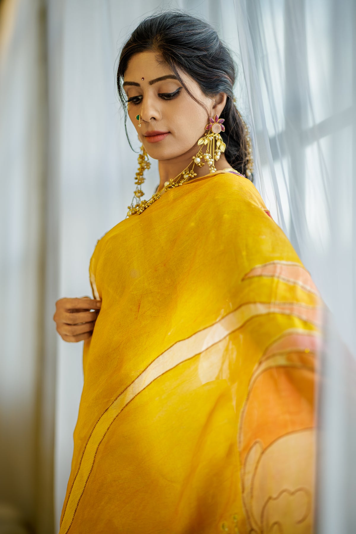 A woman wearing a mustard yellow saree with peach and cream painted floral details, styled with gold jewelry, standing in front of a soft-focus curtain background.