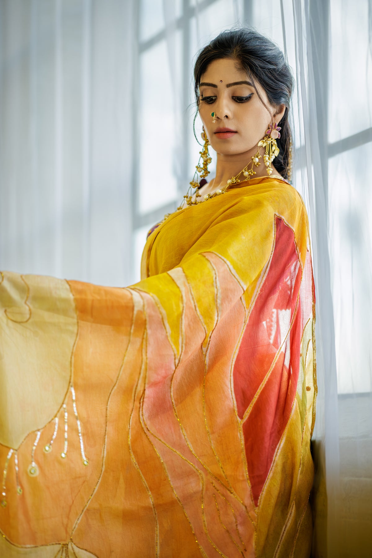 A woman wearing a yellow and orange saree with pink and peach abstract patterned borders, styled with gold jewelry, standing in front of a softly lit window with white curtains.