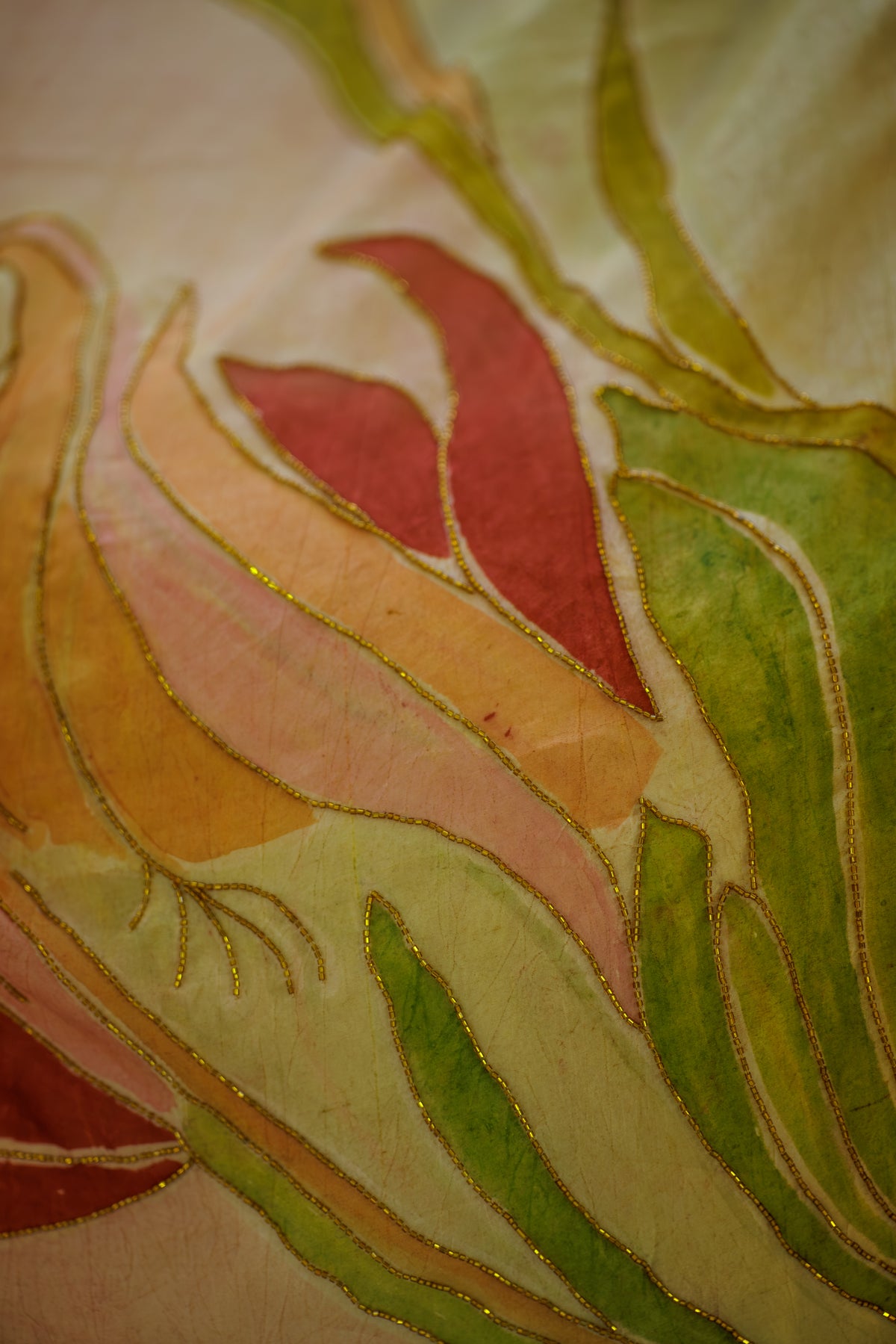 A close-up of a textile or artwork featuring abstract curved leaf-like shapes in green, yellow, peach, pink, and red outlined with metallic gold thread on a light cream background.
