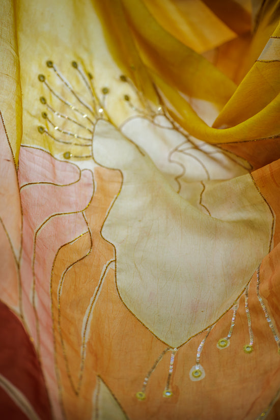 A close-up of a sheer, lightweight fabric with an abstract floral or leaf-like pattern in warm tones. The material is translucent and layered, showing shades of golden yellow, peach, soft pink, and cream, with delicate metallic embroidered details.