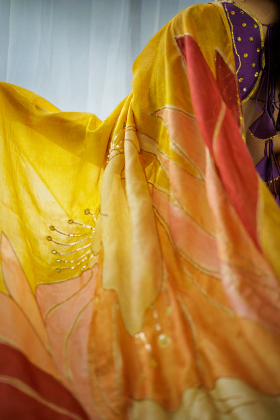 A close-up of a flowing sheer fabric with a large floral or leaf-like print in yellow, orange, peach, and red, with a small glimpse of a purple garment at the top right.