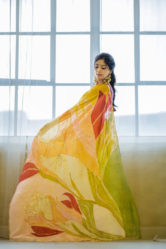 A woman wearing a large, flowing sheer scarf or shawl with a floral print in yellow, orange, green, and red, standing in front of a bright window.
