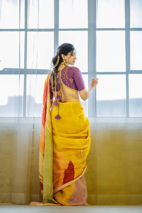 A woman wearing a yellow and orange saree with a purple embroidered blouse, standing in front of a large window with sheer curtains.